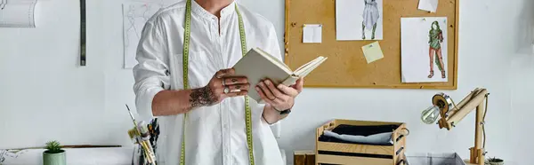 Молодой человек, владелец мастерской по реставрации одежды DIY, читает книгу в своем рабочем пространстве, окруженную инструментами и дизайном.. — стоковое фото