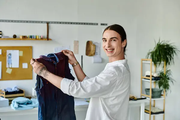 A young man in a white shirt holds up a piece of clothing hes restoring in his atelier, showcasing his commitment to sustainable fashion. — Stock Photo