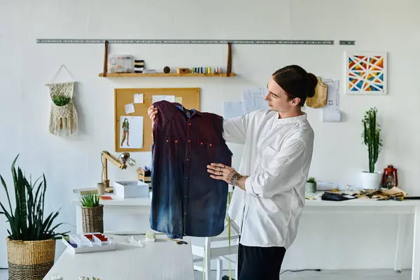 Ein junger Mann im weißen Hemd begutachtet in seinem DIY-Kleideratelier ein Vintage-Hemd und zeigt seine Leidenschaft für Nachhaltigkeit und haucht ausrangierten Kleidungsstücken neues Leben ein. — Stockfoto