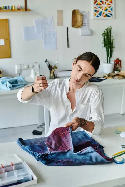 A young man in a white shirt examines a pair of upcycled jeans in his DIY clothing restoration atelier. — Stock Photo