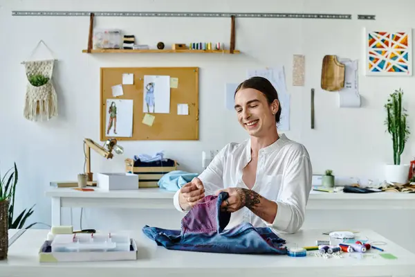 A young man smiles as he meticulously sews a piece of clothing in his brightly lit atelier. Focused on sustainability, he gives new life to discarded garments. — Stock Photo