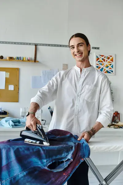 A young designer smiles as he irons a denim shirt in his clothing restoration atelier, giving new life to old items. — Stock Photo