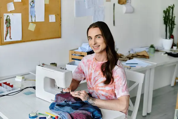 A young man, wearing a tie-dye shirt, smiles while working on a sewing machine in his clothing restoration atelier. — Stock Photo