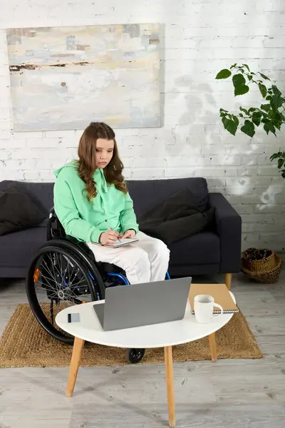 A young brunette woman sits in a wheelchair and uses a laptop in her living room. She is wearing a green sweater and white pants. — Stock Photo