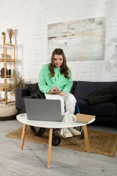A young brunette woman in a wheelchair is sitting in her living room and working on her laptop. She has a notebook and pen in her lap. — Stock Photo