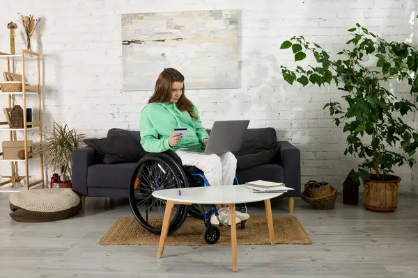 A brunette woman in a wheelchair sits in her living room, using a laptop and holding a credit card. She is browsing online while enjoying her time at home. — Stock Photo