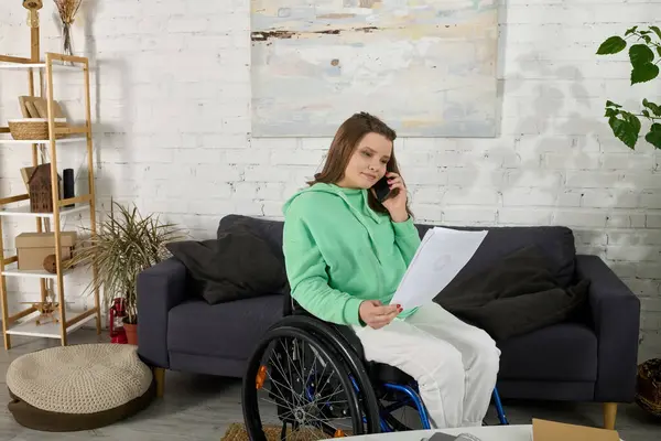 Une jeune femme aux cheveux bruns s'assoit dans un fauteuil roulant, examinant la paperasse lors d'un appel téléphonique dans son salon. — Photo de stock