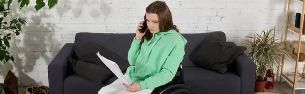 A young woman with brown hair sits in a wheelchair, talking on the phone while holding papers. Shes in her living room, surrounded by home furnishings. — Stock Photo