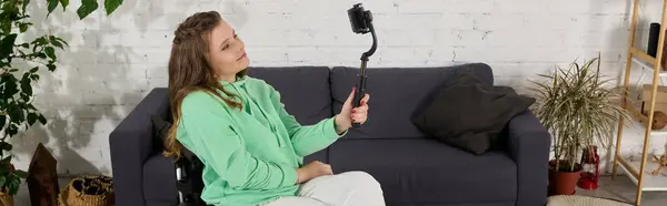 Uma jovem morena em uma cadeira de rodas se senta em sua sala de estar e segura um smartphone para gravar um vídeo. — Fotografia de Stock