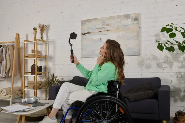 A young brunette woman sits in a wheelchair in her living room, using a selfie stick to film a video. — Stock Photo
