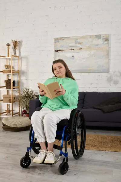 A young brunette woman sits in a wheelchair in her living room, engrossed in reading a book. — Stock Photo