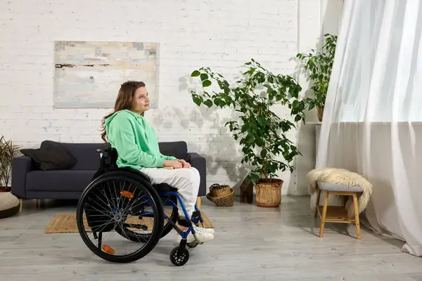 A young woman with brown hair sits in a wheelchair in a living room. She looks out the window, appearing thoughtful. — Stock Photo