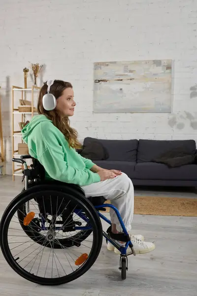 Une jeune femme aux cheveux bruns est assise dans un fauteuil roulant dans un salon, portant un casque et écoutant de la musique. — Stock Photo