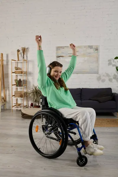 A young brunette woman with a disability sits in a wheelchair in her living room and dances along to music playing in her headphones. — Stock Photo