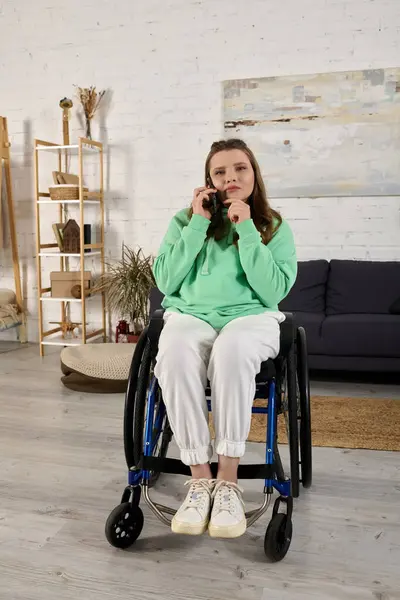 A young woman in a wheelchair sits in her living room, listening intently to a phone call. — Stock Photo