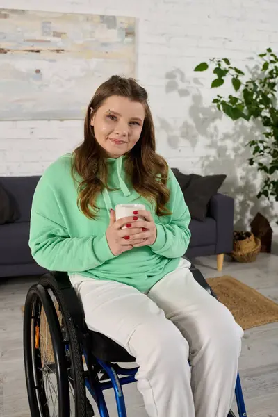 A brunette woman in a wheelchair smiles as she holds a mug in her living room. — Stock Photo