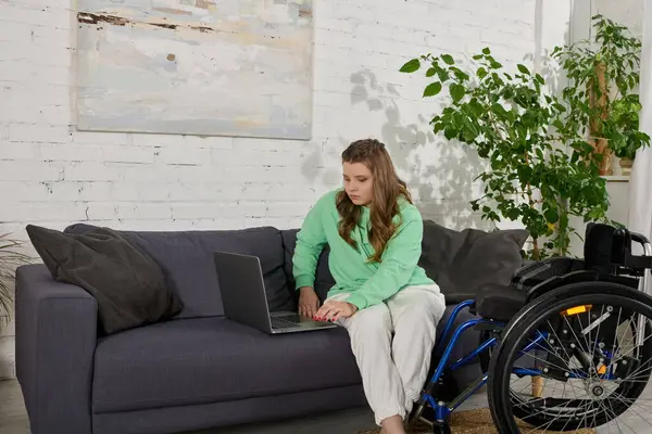 Eine junge brünette Frau mit Behinderung sitzt auf einer Couch in ihrem Wohnzimmer und benutzt einen Laptop. Ihr Rollstuhl steht neben ihr. — Stockfoto