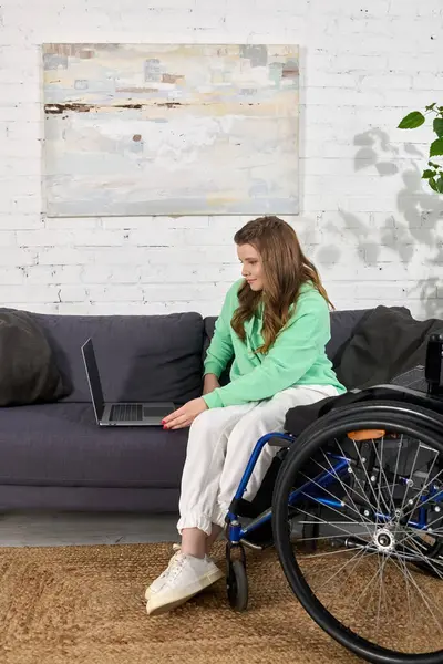 A young brunette woman in a wheelchair is using a laptop while sitting in her living room. — Stock Photo
