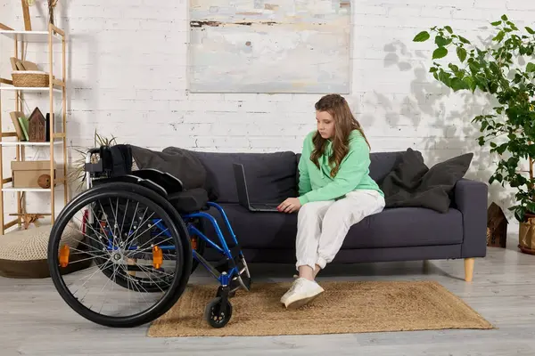 Une jeune femme handicapée s'assoit sur un canapé dans son salon, à l'aide d'un ordinateur portable. — Photo de stock