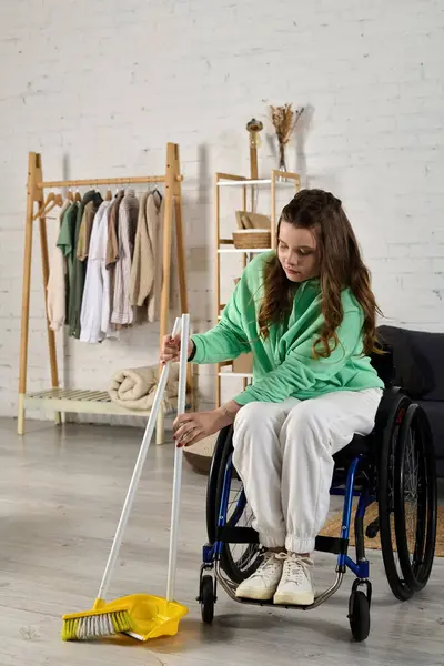 A young woman in a wheelchair sits in her living room, sweeping the floor with a broom and dustpan. — Stock Photo