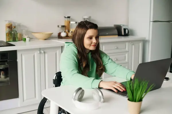 Una joven morena se sienta en una silla de ruedas y trabaja en un portátil en la mesa de su cocina. — Stock Photo