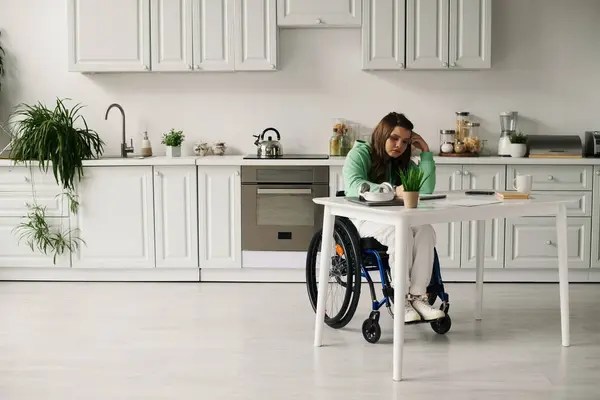 A young brunette woman in a wheelchair sits at a kitchen table, looking down at something in her lap. — Stock Photo