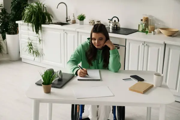 Eine junge Frau im Rollstuhl sitzt an einem Tisch in ihrem Haus und schreibt in ein Notizbuch. — Stockfoto