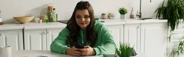 Eine junge brünette Frau in grünem Kapuzenpulli sitzt in ihrem Wohnzimmer und benutzt ihr Handy, ihr Lächeln deutet darauf hin, dass sie gut gelaunt ist. — Stockfoto