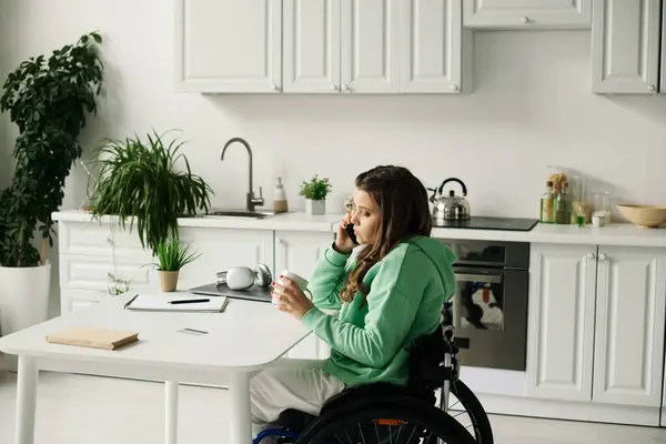 A young brunette woman in a wheelchair sits at a table in her kitchen, talking on her phone while holding a mug. — Stock Photo