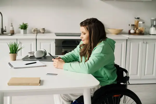 Eine brünette Frau sitzt zu Hause im Rollstuhl und benutzt ihr Handy, während sie an einem Tisch arbeitet. — Stock Photo