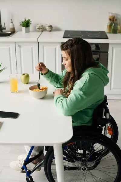 A young brunette woman in a wheelchair sits at a table in her living room, enjoying a bowl of cereal and a glass of orange juice. — Stock Photo