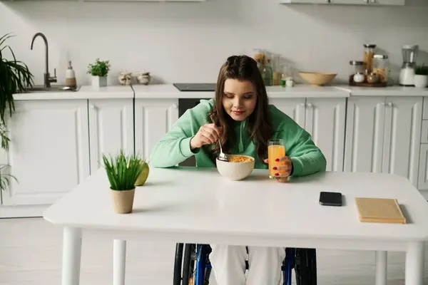 Uma jovem mulher em uma cadeira de rodas se senta em uma mesa de cozinha comendo cereais e bebendo suco de laranja. — Fotografia de Stock