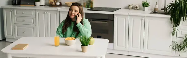 A young brunette woman in a wheelchair sits at a table in her kitchen, talking on the phone. — Stock Photo