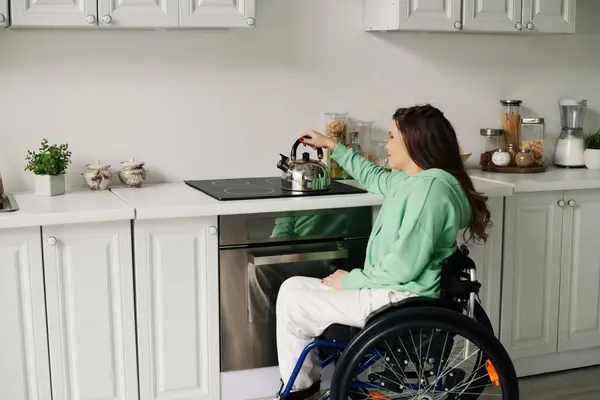 Une brune en fauteuil roulant s'assoit dans sa cuisine, cherchant à mettre une bouilloire sur la cuisinière. — Photo de stock