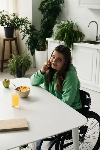 A young brunette woman with a disability sits in a wheelchair at her kitchen table, talking on her cell phone. — Stock Photo