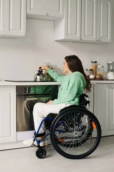 Une jeune femme aux cheveux bruns s'assoit dans un fauteuil roulant dans sa cuisine, cherchant une bouilloire sur la cuisinière. — Photo de stock