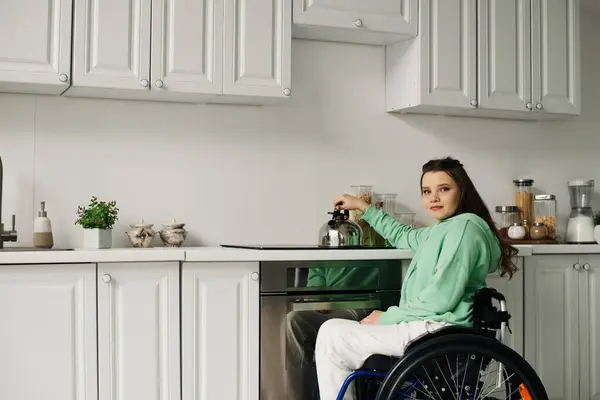 A young brunette woman in a wheelchair is preparing dinner in her kitchen. She is wearing a green sweatshirt and white pants. — Stock Photo
