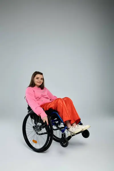 A brunette young woman in a pink sweatshirt and orange pants smiles while sitting in a wheelchair in a studio setting. — Stock Photo