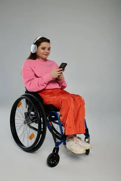 A young woman in a wheelchair wears headphones and checks her phone. — Stock Photo
