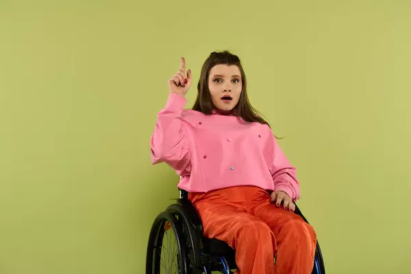 A young woman in a wheelchair sits in a studio setting, pointing upwards with a surprised expression on her face. — Stock Photo