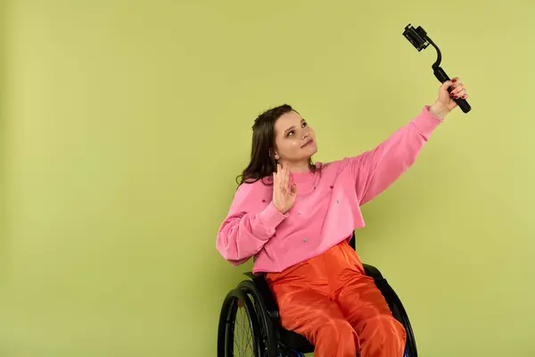Una giovane donna bruna su una sedia a rotelle, che indossa abiti casual, sorride mentre si fa un selfie con un bastone di selfie in un ambiente da studio. — Foto stock