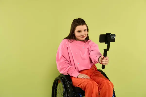 A young brunette woman with a disability is seated in a wheelchair in a studio setting. She is wearing casual attire and smiling as she films a video with a phone on a selfie stick. — Stock Photo