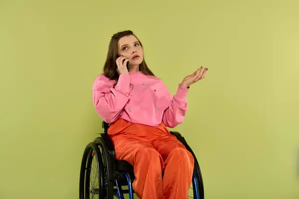 Una joven con el pelo castaño largo se sienta en una silla de ruedas en un estudio, hablando por teléfono mientras hace un gesto con su mano libre. — Stock Photo