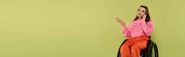 A young brunette woman with a disability sits in a wheelchair, holding a phone to her ear, in a studio setting. She is wearing casual attire. — Stock Photo