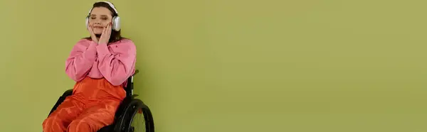 A brunette woman in a wheelchair sits in front of a green background, listening to music with headphones. — Stock Photo