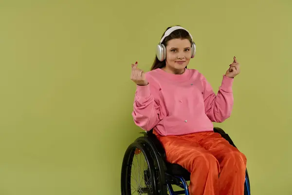 A young woman with brown hair sits in a wheelchair in a studio setting, wearing a pink sweater and orange pants. She wears headphones and smiles as she listens to music. — Stock Photo
