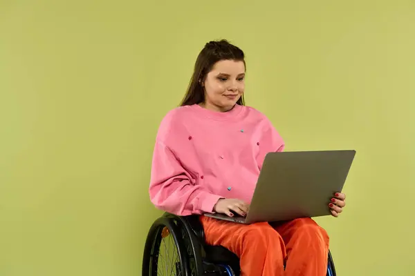 A young woman with brown hair sits in a wheelchair in a studio setting, using a laptop. — Stock Photo