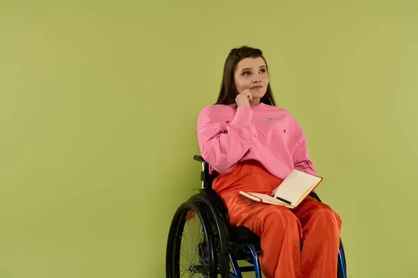 Une femme brune est assise dans un fauteuil roulant dans un studio, regardant doucement sur le côté. Elle est vêtue occasionnellement d'un pull rose et d'un pantalon orange. — Photo de stock