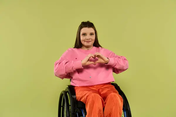 A young brunette woman in a wheelchair smiles while making a heart shape with her hands in front of a green background. — Stock Photo
