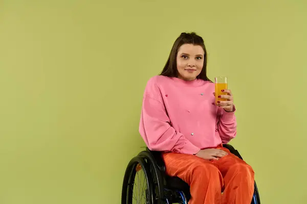 Una joven morena con discapacidad se sienta en una silla de ruedas en un estudio, casualmente vestida y sosteniendo un vaso de jugo. — Stock Photo